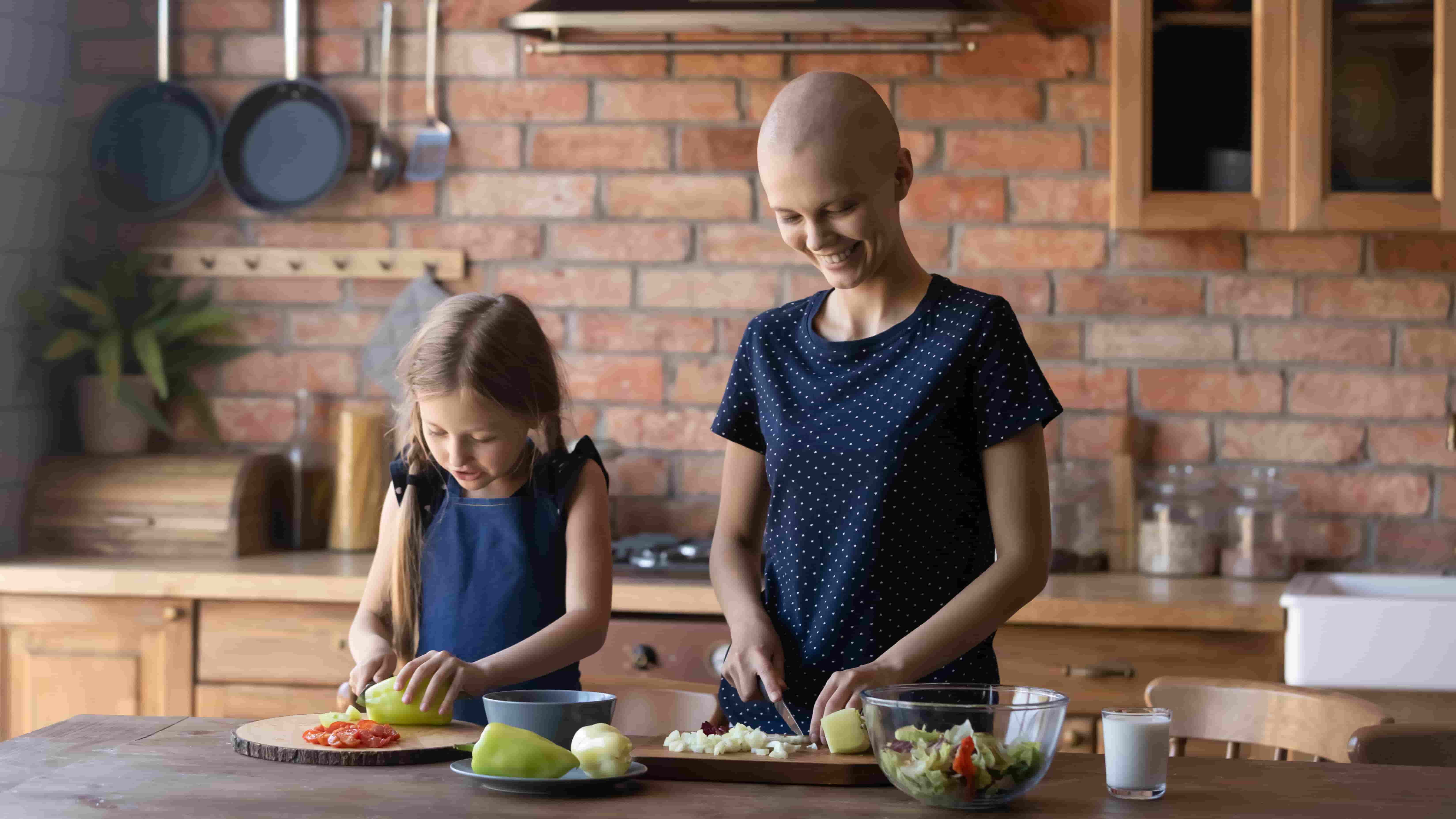 Mother with a bald head from cancer treatment cuts fruit with her daughter in a kitchen
