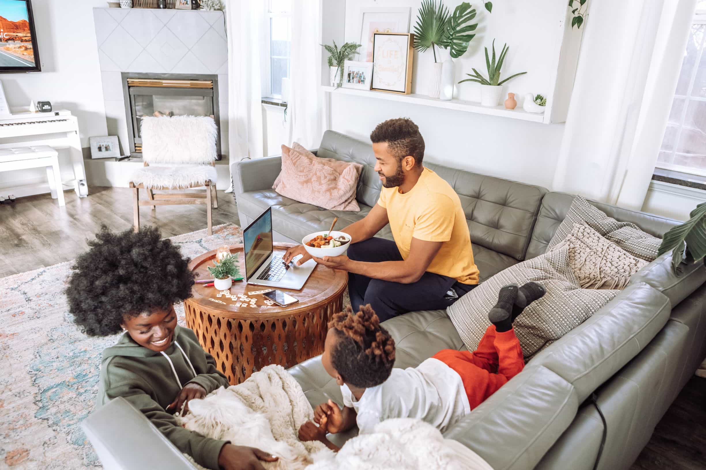 A mother, father and their child lounge on a corner sofa whilst watching television and eating